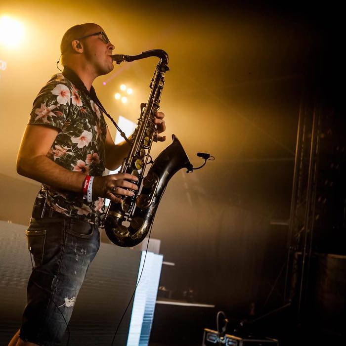 Sakso speelt live saxofoon te midden van een feestende menigte, met kleurrijke lichteffecten en opzwepende sfeer.