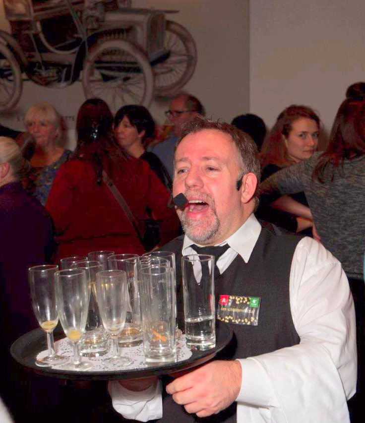 De Zingende Ober zingt tussen feestende gasten terwijl hij drankjes serveert met een glimlach.
